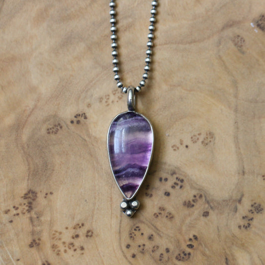 May include: A teardrop-shaped pendant necklace featuring a polished, purple and white stone. The stone is encased in a silver-colored metal setting and hangs from a delicate, beaded chain. The pendant has a small, three-dot design at the bottom.