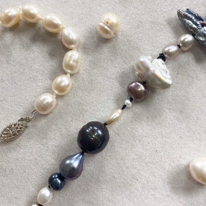A close-up of a beaded necklace laid on a light beige textured surface. The necklace features a mix of creamy white baroque pearls, dark navy and gray teardrop beads, small lavender-gray beads, and a silver-toned leaf-shaped charm.