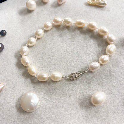 A freshwater pearl bracelet with irregular baroque pearls and a silver-toned leaf clasp, displayed on a light gray textured surface with loose pearls nearby.