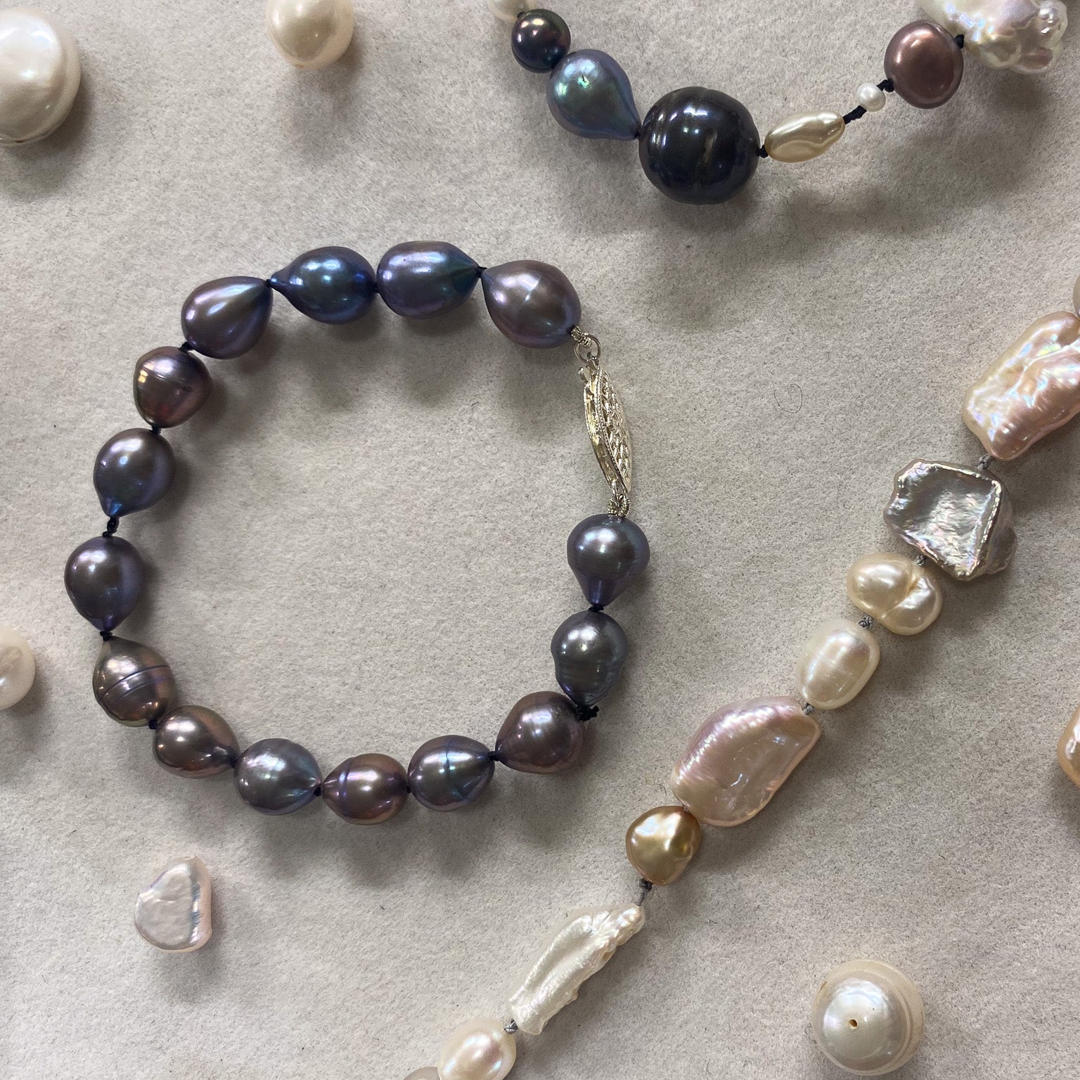 A jewelry flat-lay featuring a dark gray baroque pearl bracelet and a long mixed-bead necklace with baroque pearls in cream, pink, and gray, arranged on a textured beige surface.