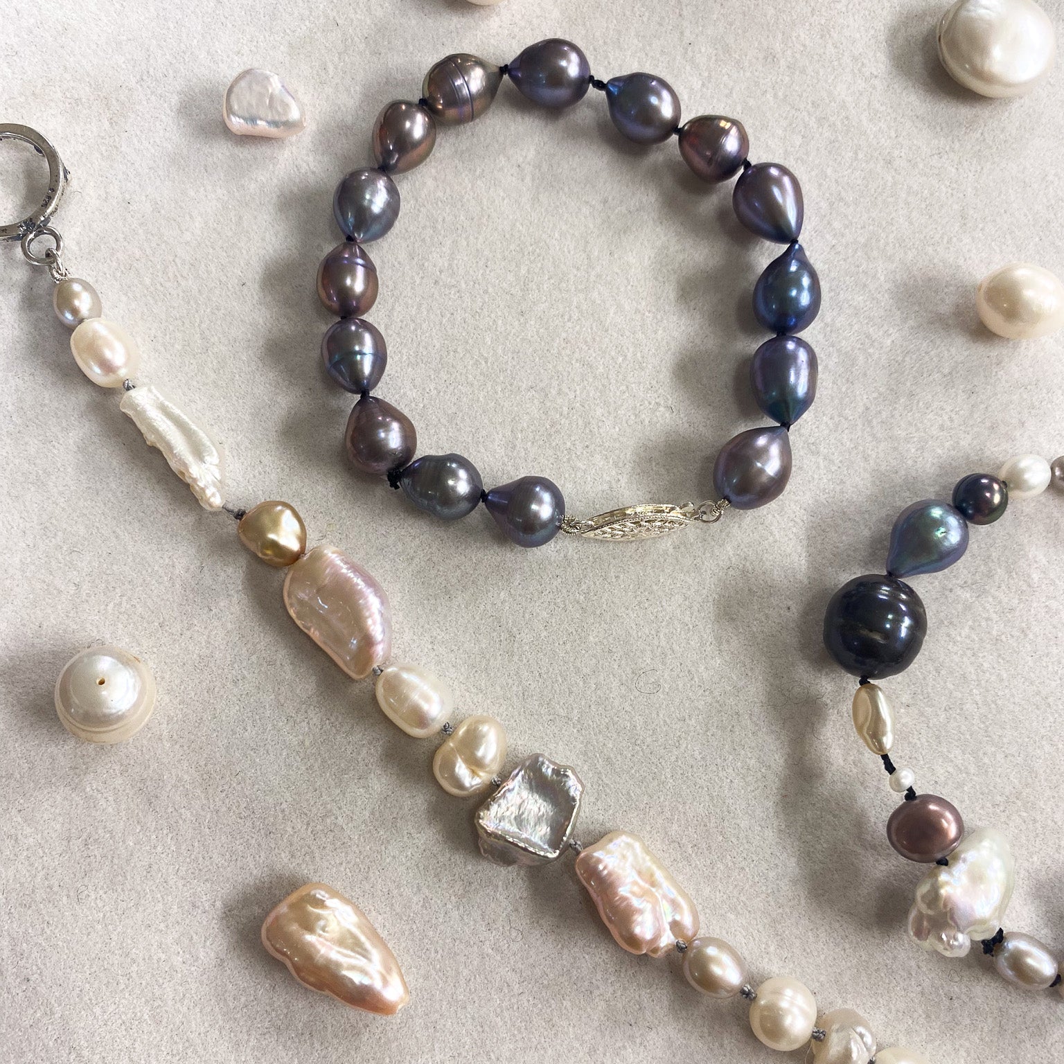 A jewelry set featuring a beaded bracelet and matching beaded necklace made of baroque pearls in shades of cream, beige, and dark gray, displayed on a light beige textured surface.