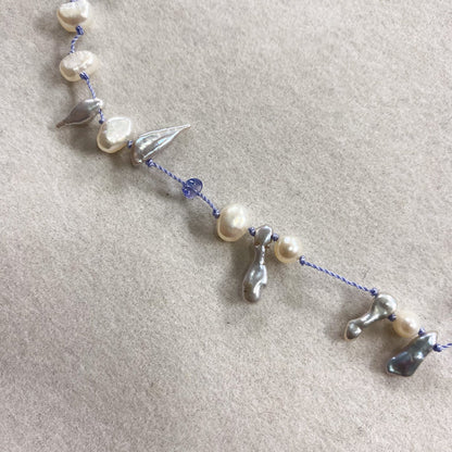 A delicate beaded necklace featuring irregular white baroque pearls, small silver metallic charms, and a blue cord, laid out on a textured beige fabric background.