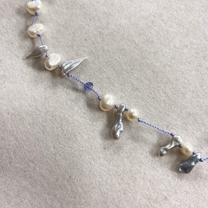 A delicate beaded necklace featuring irregular white baroque pearls, small silver metallic charms, and a blue cord, laid out on a textured beige fabric background.