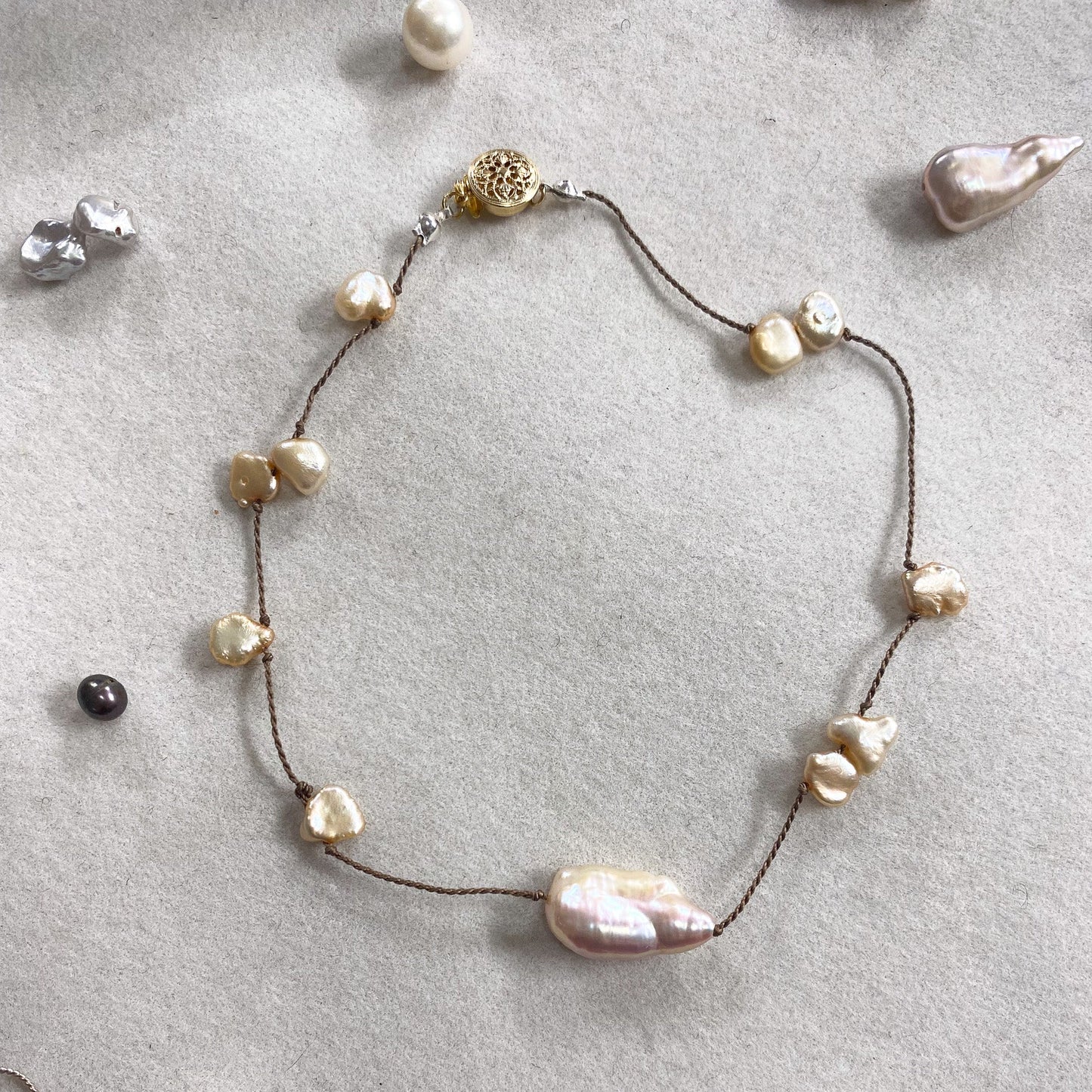 A delicate necklace with irregular baroque pearls on a dark cord, displayed on a textured light gray surface with other jewelry pieces scattered around.