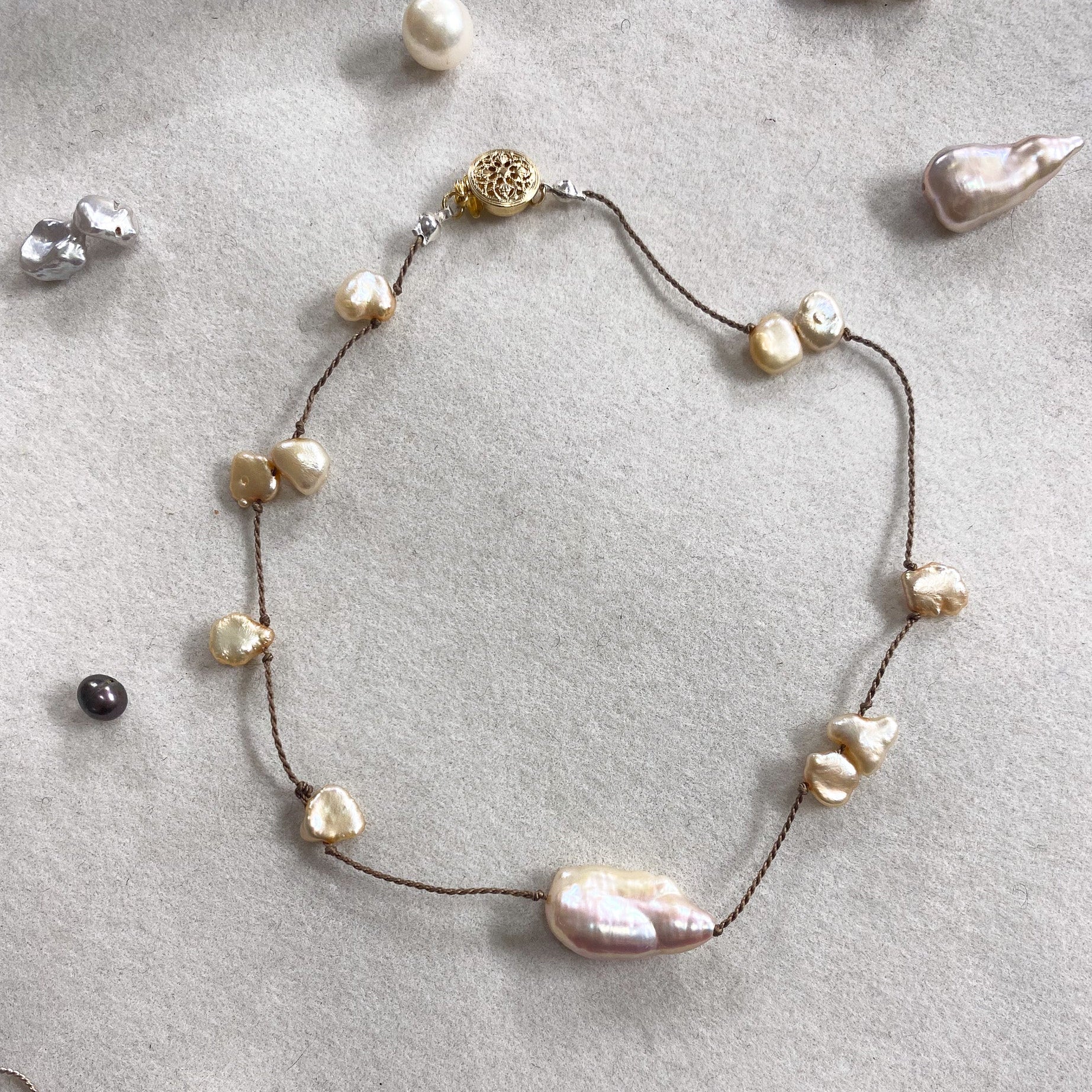 A delicate necklace with irregular baroque pearls on a dark cord, displayed on a textured light gray surface with other jewelry pieces scattered around.