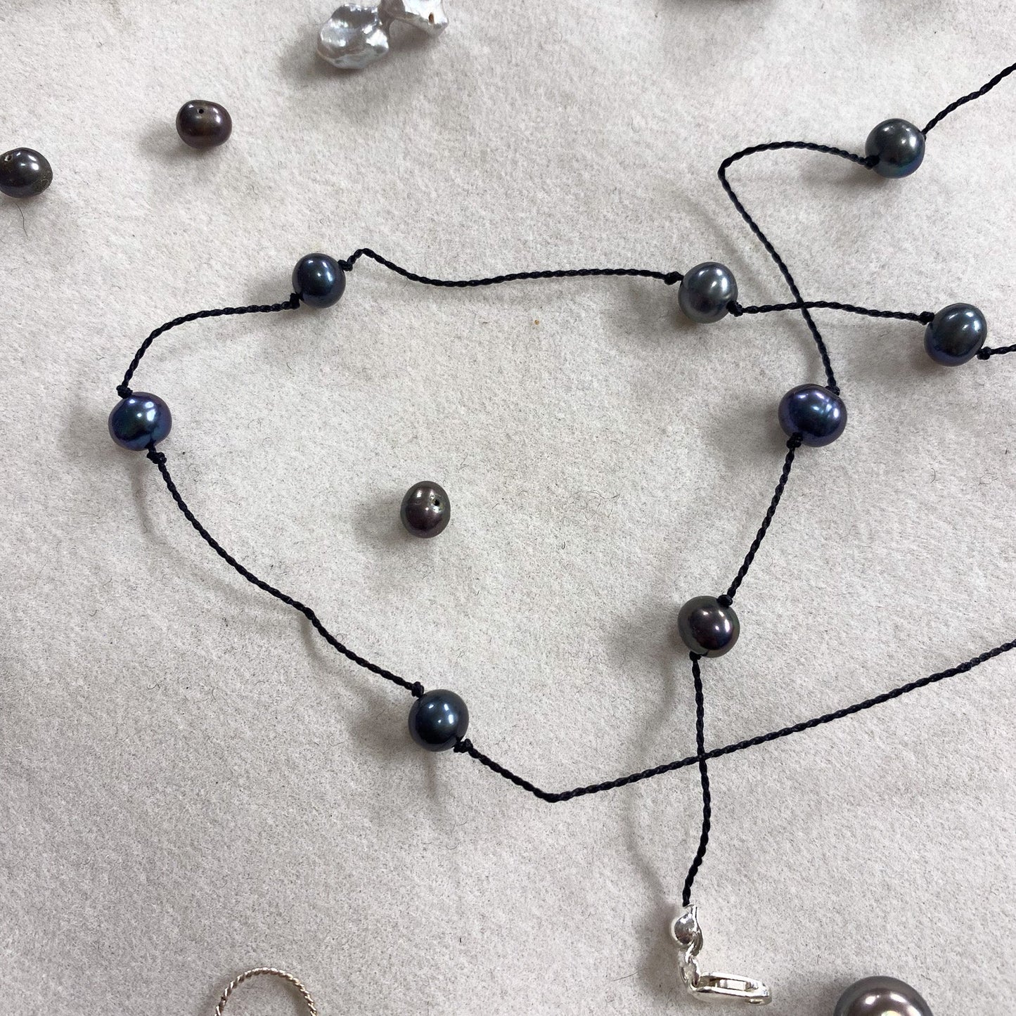 A black cord necklace with dark blue-gray round pearls and a small silver charm, laid out on a light gray textured surface with other jewelry pieces nearby.