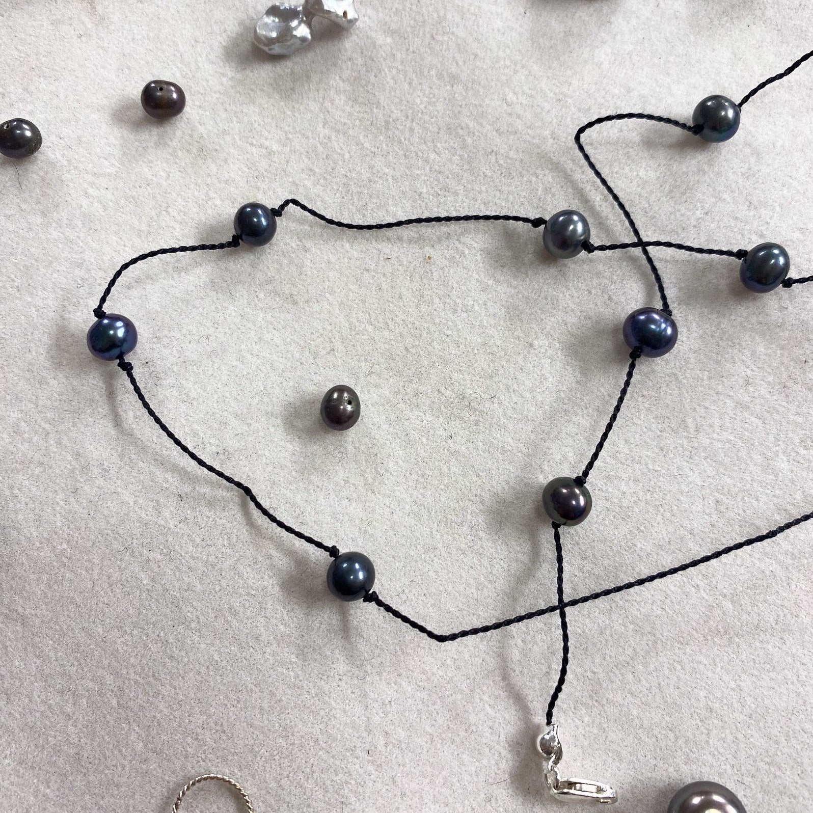 A black cord necklace with dark blue-gray round pearls and a small silver charm, laid out on a light gray textured surface with other jewelry pieces nearby.