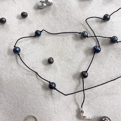A black cord necklace with dark blue-gray round pearls and a small silver charm, laid out on a light gray textured surface with other jewelry pieces nearby.