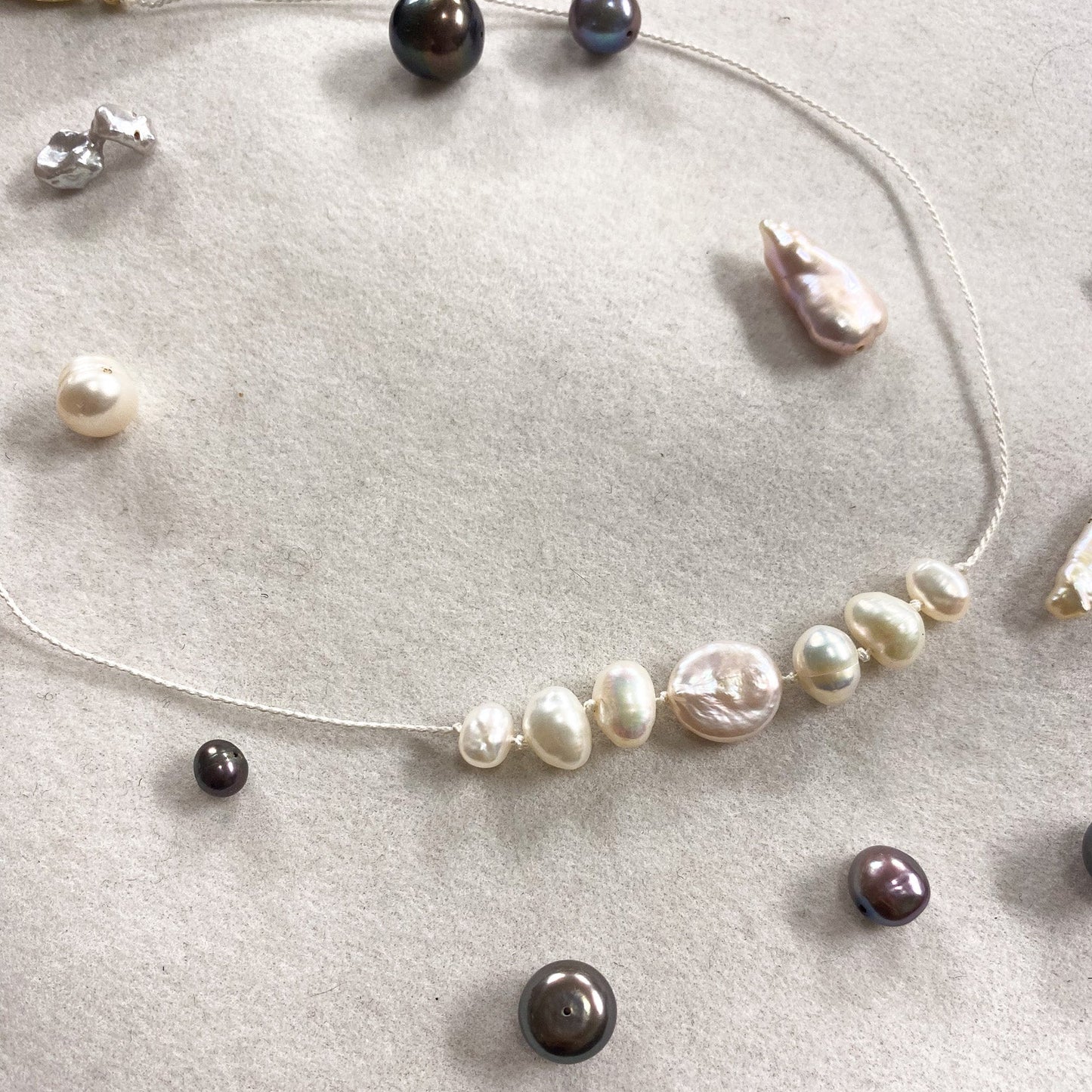 A delicate beaded necklace with irregular baroque pearls in white, cream, and dark brown tones, displayed on a textured light beige surface with other jewelry pieces scattered around.