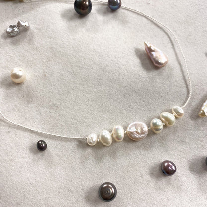 A delicate beaded necklace with irregular baroque pearls in white, cream, and dark brown tones, displayed on a textured light beige surface with other jewelry pieces scattered around.