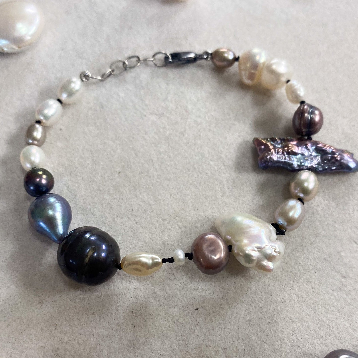 A handmade baroque pearl beaded bracelet with a silver clasp, displayed on a neutral beige textured surface with other pearls scattered around.