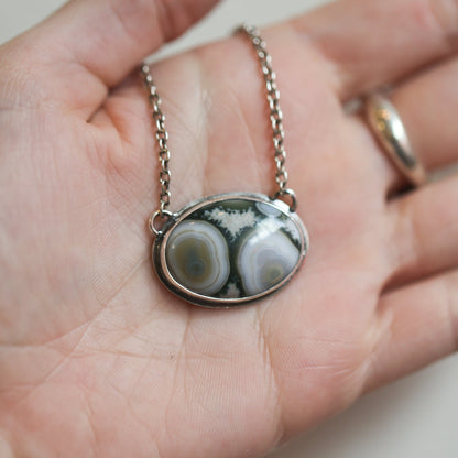 May include: A silver chain necklace with an oval pendant featuring a green and white agate stone with a swirling pattern.