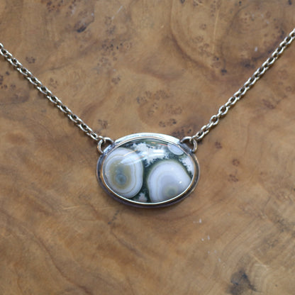 May include: A silver chain necklace with a silver oval pendant featuring a green and white agate stone with a swirling pattern.