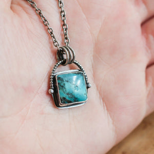 May include: A silver necklace with a square turquoise stone pendant. The pendant is set in a silver frame and has a swiveling design.
