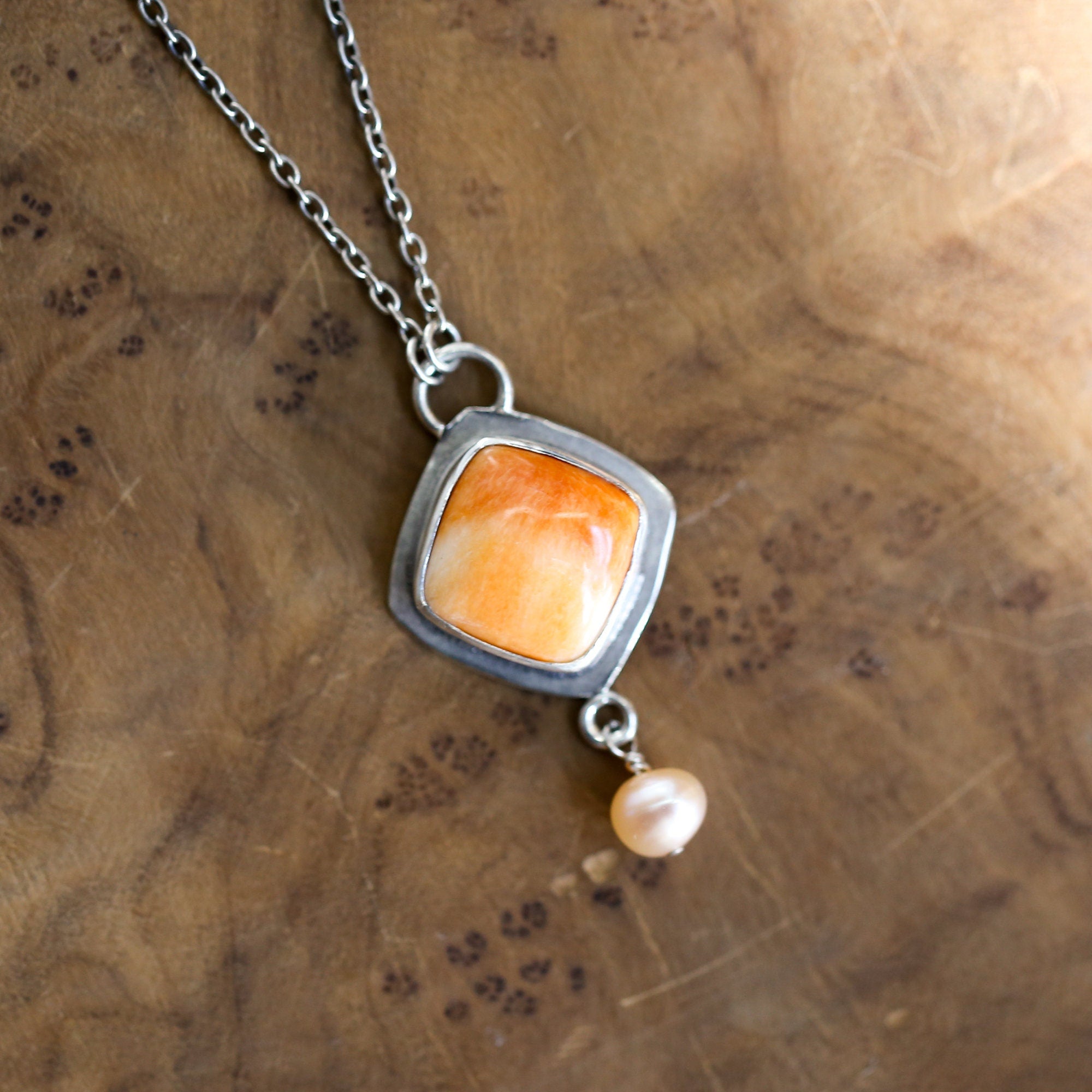 May include: A silver necklace with a square pendant featuring an orange gemstone and a single pearl drop.