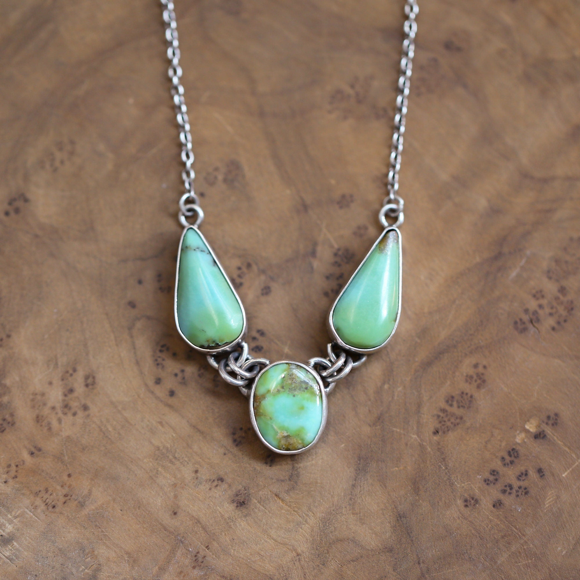 May include: A silver necklace with three turquoise stones. The stones are in the shape of teardrops and are set in silver. The necklace is on a wooden surface.