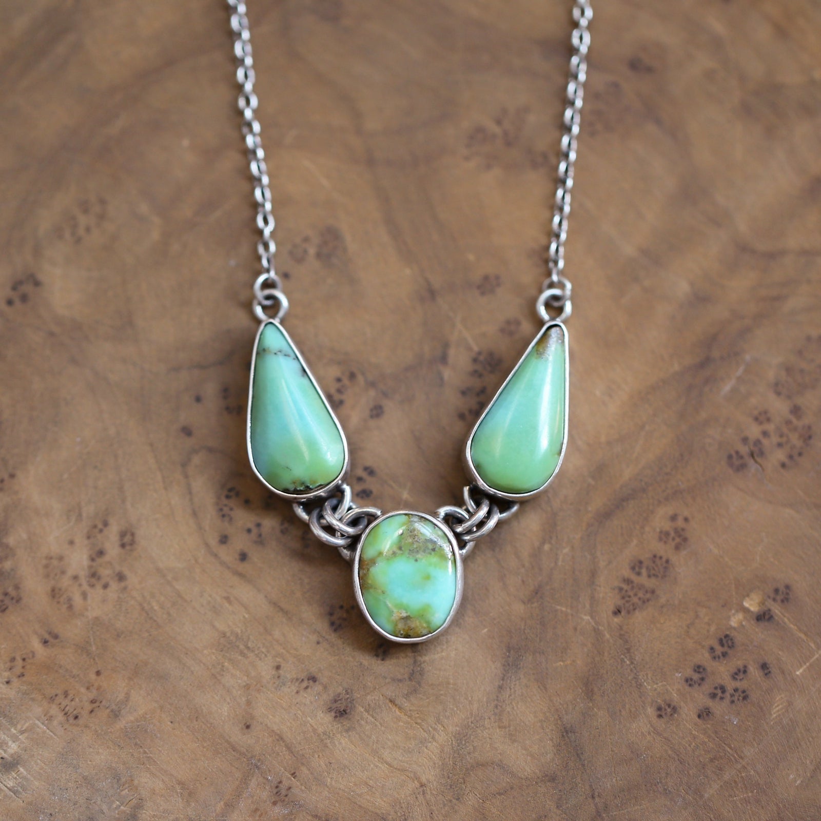 May include: A silver necklace with three turquoise stones. The stones are in the shape of teardrops and are set in silver. The necklace is on a wooden surface.