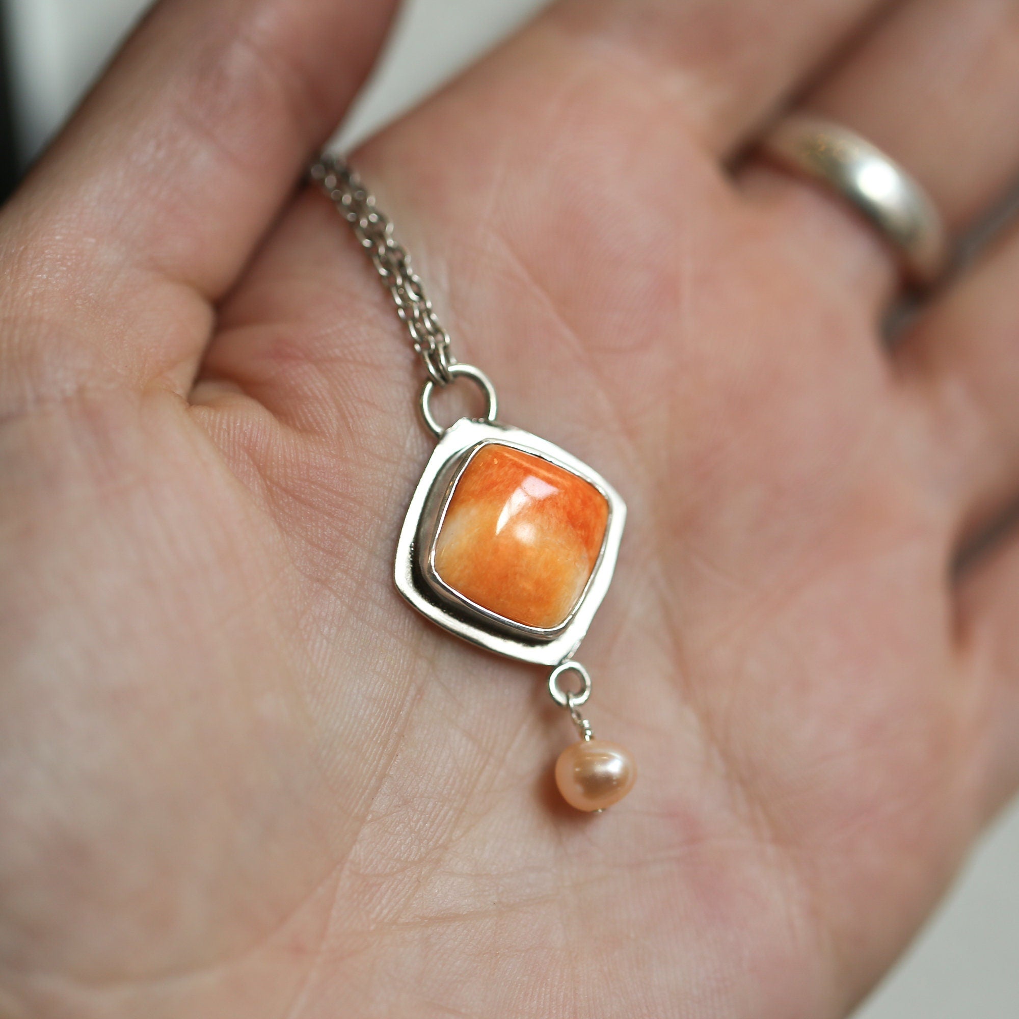 May include: A silver necklace with a square pendant featuring an orange and white gemstone. The pendant has a small pearl charm hanging from the bottom.