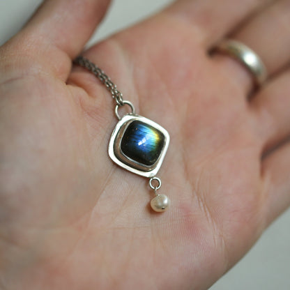 May include: A silver pendant necklace with a square-shaped labradorite gemstone and a single pearl drop. The labradorite gemstone has a blue, green, and gold iridescent sheen.
