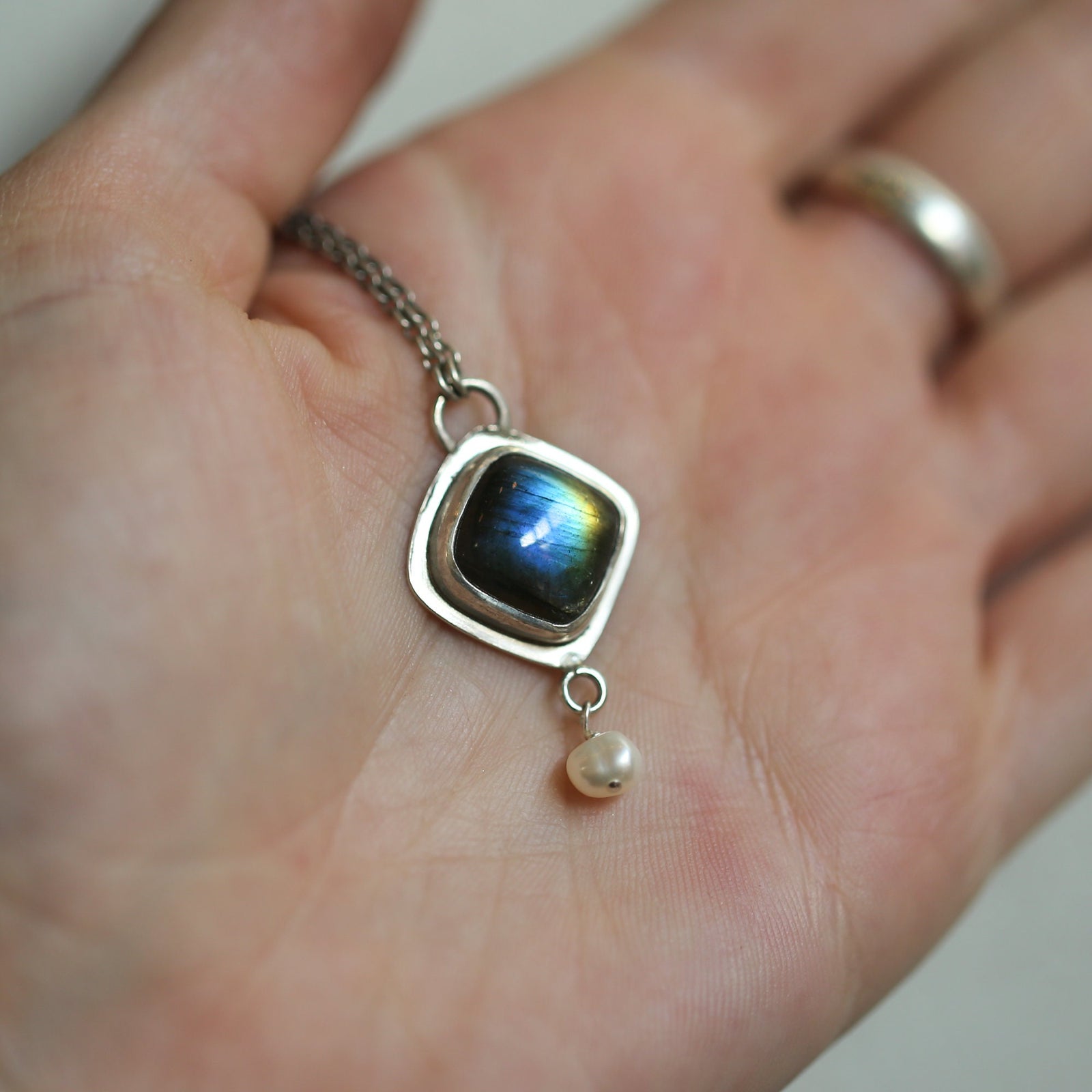 May include: A silver pendant necklace with a square-shaped labradorite gemstone and a single pearl drop. The labradorite gemstone has a blue, green, and gold iridescent sheen.