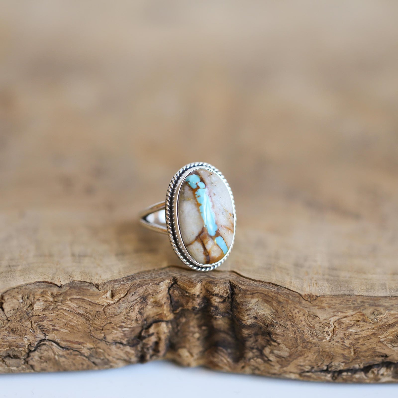 May include: Oval-shaped sterling silver ring featuring a unique turquoise gemstone. The stone showcases a blend of creamy beige, brown, and light blue hues, creating a captivating natural pattern. The ring's band is delicately crafted with a twisted rope design, adding a touch of elegance. This handcrafted piece is a statement of natural beauty and exquisite craftsmanship.