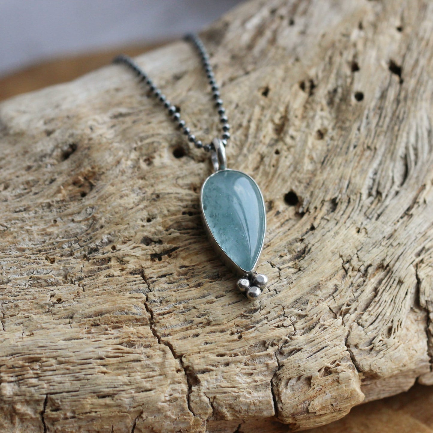 May include: A silver chain necklace with a teardrop-shaped aquamarine pendant. The pendant is set in silver and has a beaded detail at the bottom.