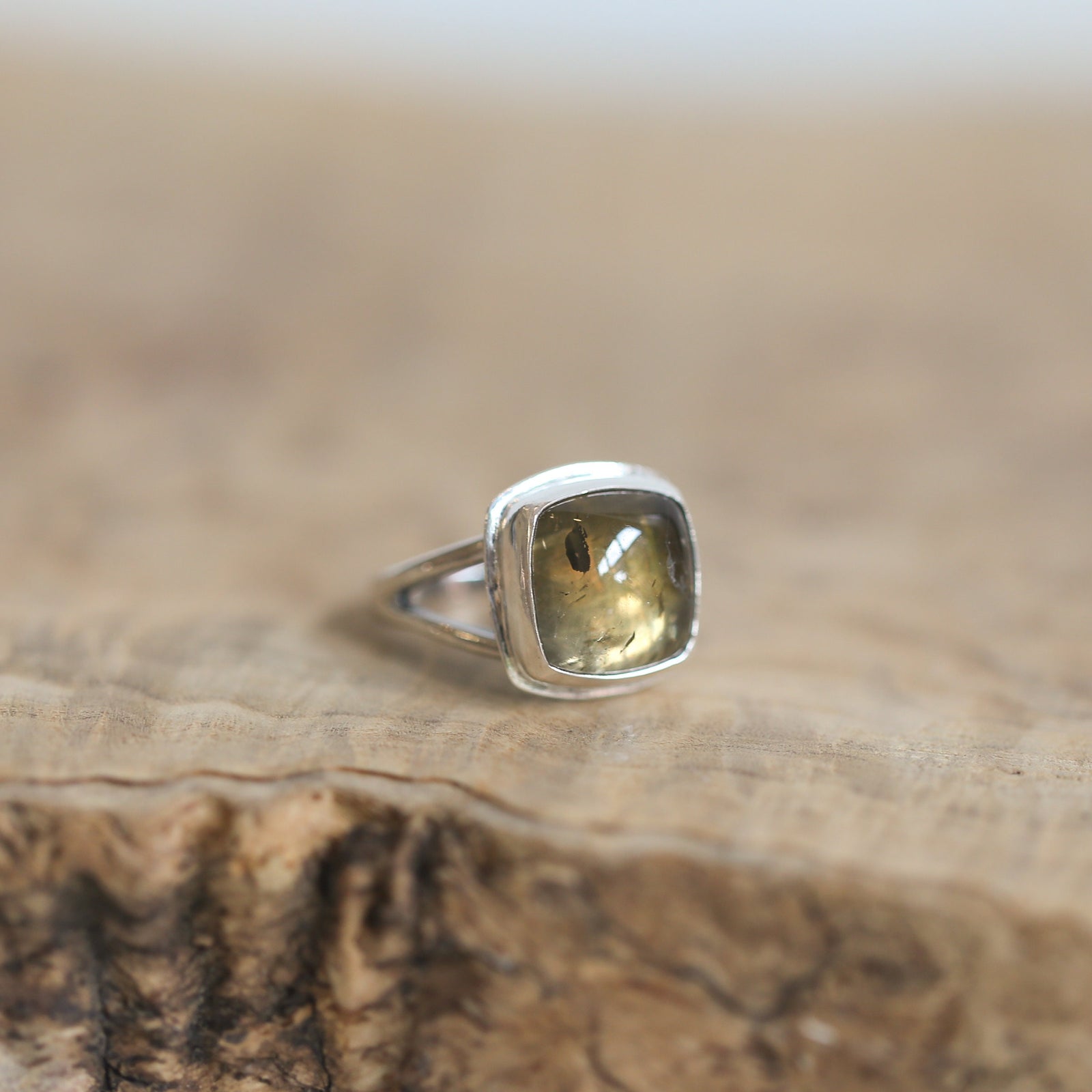 May include: Silver ring featuring a cushion-cut, translucent greenish-brown gemstone. The stone has subtle darker inclusions visible within its structure. The ring's setting is a simple, elegant design with two delicate prongs holding the gemstone securely. The overall style is understated and sophisticated, suitable for everyday wear or special occasions. The gemstone's unique color and inclusions add a touch of natural beauty to the piece.