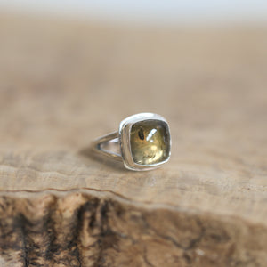 May include: Silver ring featuring a cushion-cut, translucent greenish-brown gemstone. The stone has subtle darker inclusions visible within its structure. The ring's setting is a simple, elegant design with two delicate prongs holding the gemstone securely. The overall style is understated and sophisticated, suitable for everyday wear or special occasions. The gemstone's unique color and inclusions add a touch of natural beauty to the piece.