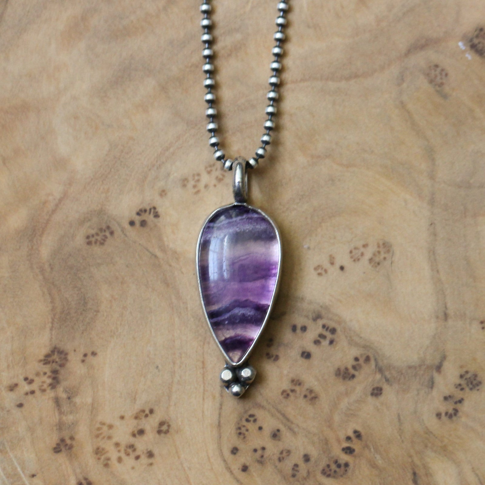 May include: A teardrop-shaped pendant necklace featuring a polished, purple and white stone. The stone is encased in a silver-colored metal setting and hangs from a delicate, beaded chain. The pendant has a small, three-dot design at the bottom.