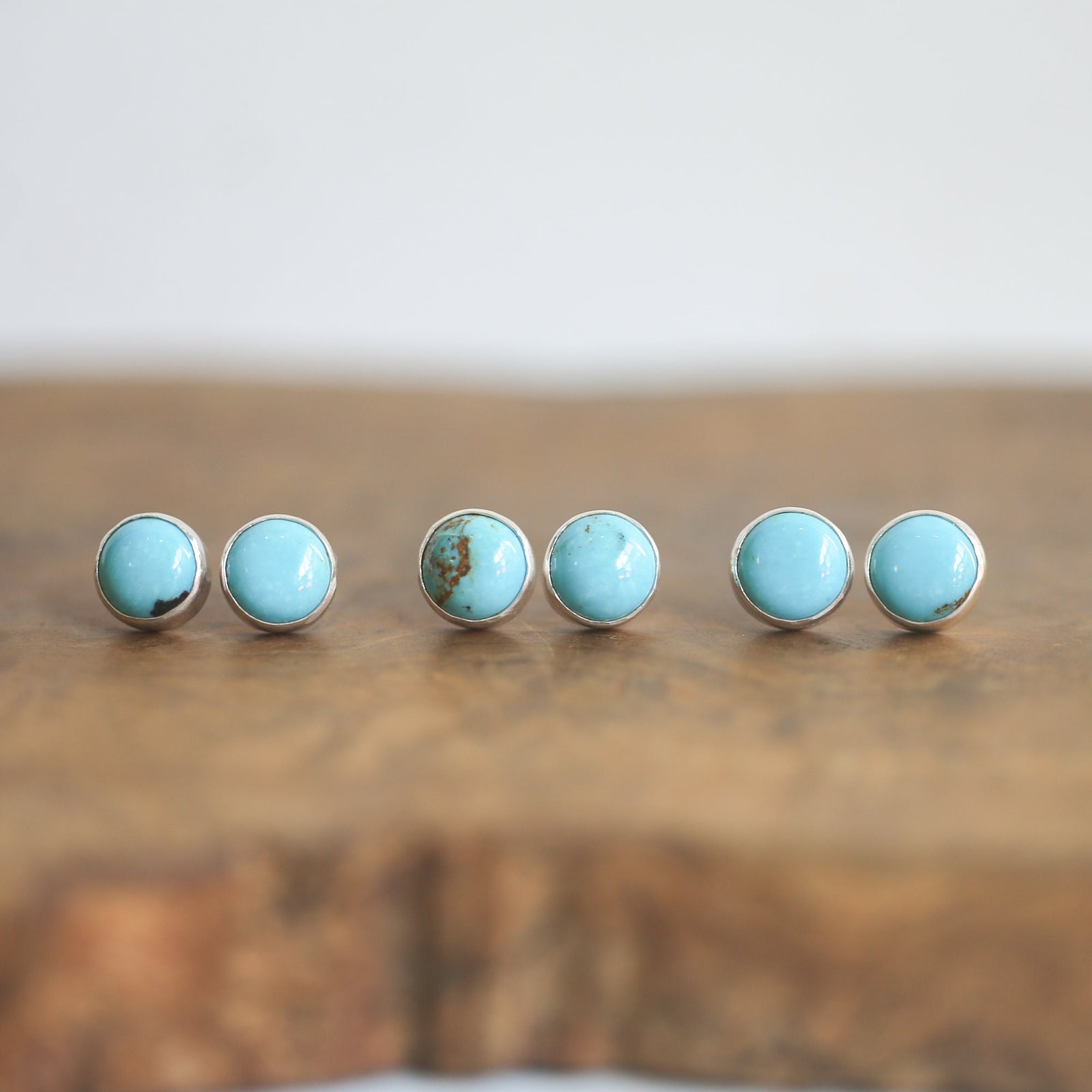 May include: Three pairs of turquoise stud earrings with silver settings. The earrings are arranged in a row on a wooden surface.