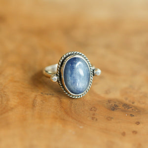 May include: A silver ring with a blue oval gemstone set in a silver bezel. The ring has a rope-like design around the bezel and small silver accents on either side of the stone.