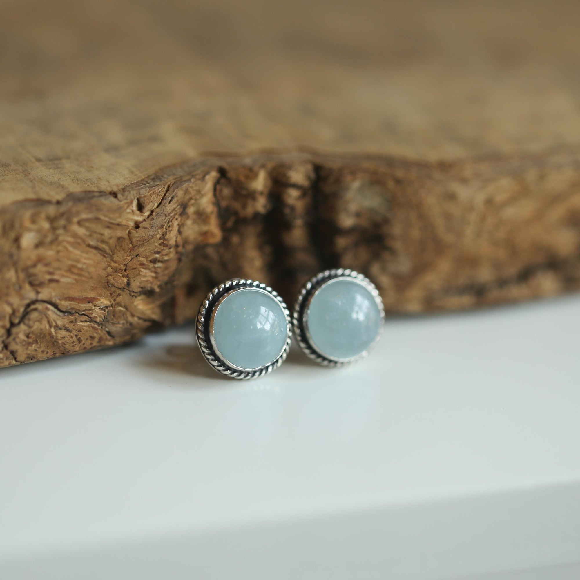 May include: A pair of stud earrings with a round, light blue gemstone center. The gemstones are set in a silver-colored metal with a twisted rope design around the edge. The earrings are displayed on a white surface with a blurred wooden background.