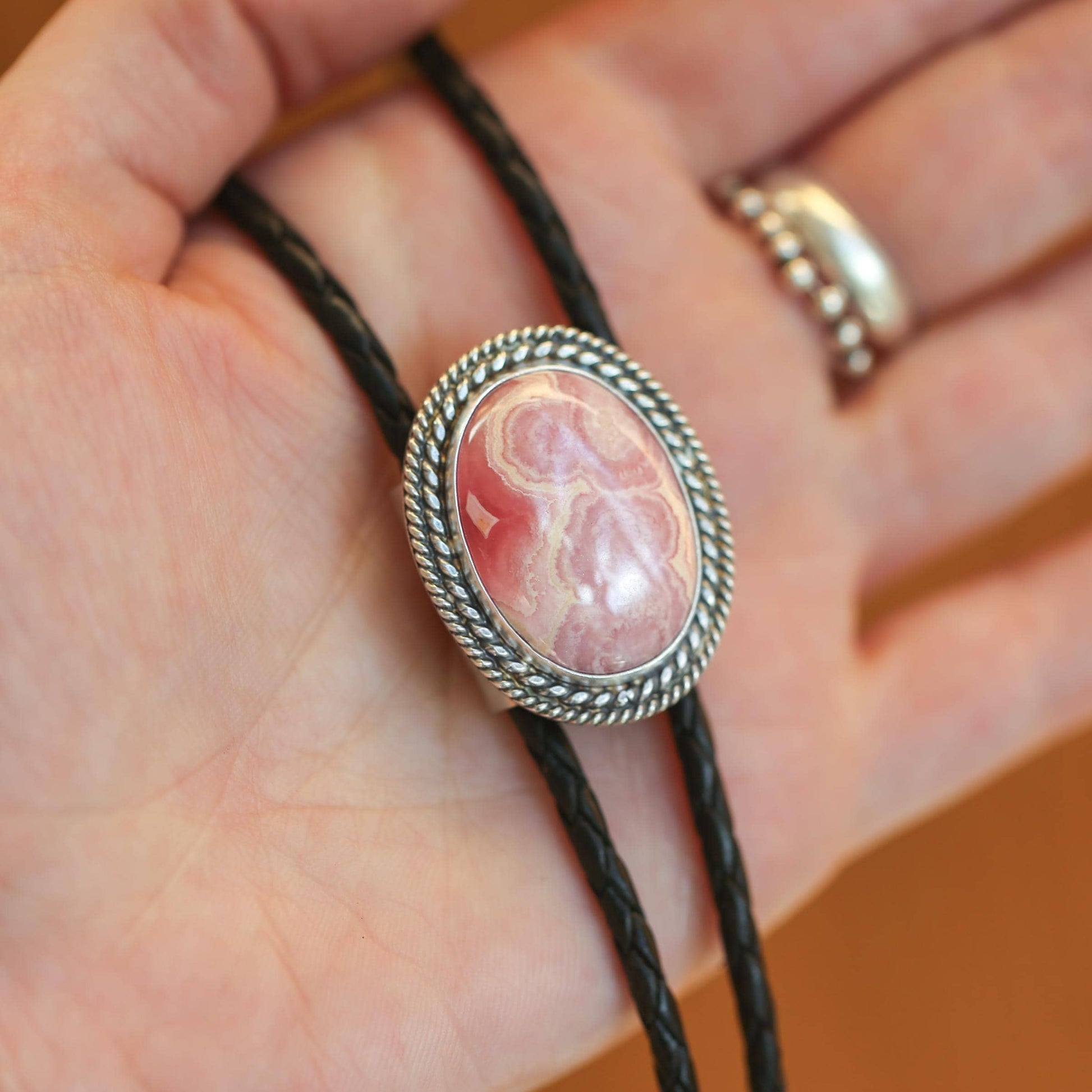May include: Boho style pink rhodonite gemstone bolo tie.  The oval shaped stone is set in a silver tone rope-edged bezel.  The bolo features black braided leather cords.  This southwestern style necklace is a unique statement piece.
