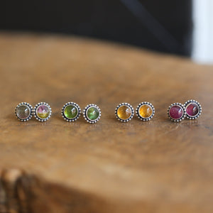 May include: Four pairs of silver stud earrings with different colored gemstones. The earrings are arranged in a row on a wooden surface. The gemstones are green, pink, yellow, and a mix of pink and green.