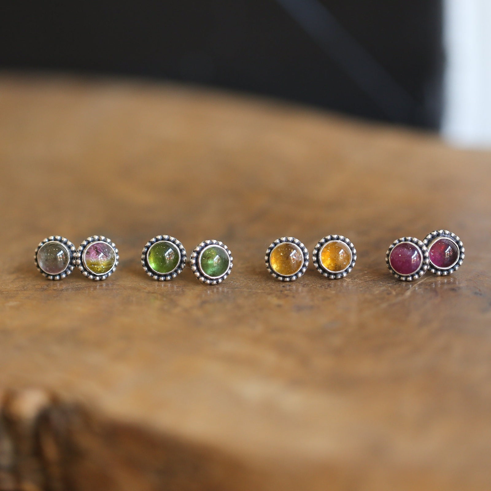 May include: Four pairs of silver stud earrings with different colored gemstones. The earrings are arranged in a row on a wooden surface. The gemstones are green, pink, yellow, and a mix of pink and green.