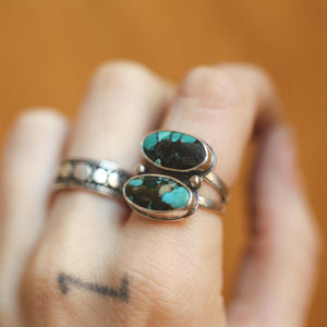 May include: A silver ring with two oval turquoise stones. The stones have a dark matrix pattern. The ring is on a finger, and another ring with a dotted design is also visible. The background is a blurred orange color.
