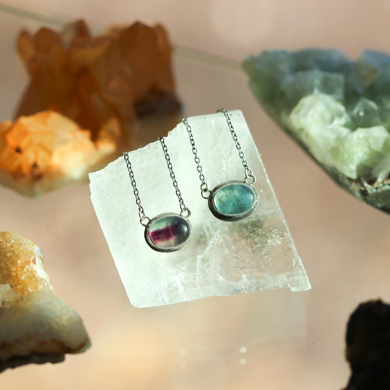 May include: Two silver necklaces with oval-shaped fluorite pendants. One pendant is a light green color and the other is a darker green with pink and purple hues. The necklaces are displayed on a white surface.