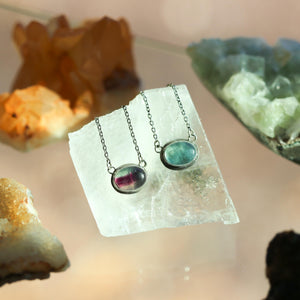 May include: Two silver necklaces with oval-shaped fluorite pendants. One pendant is a light green color and the other is a darker green with pink and purple hues. The necklaces are displayed on a white surface.
