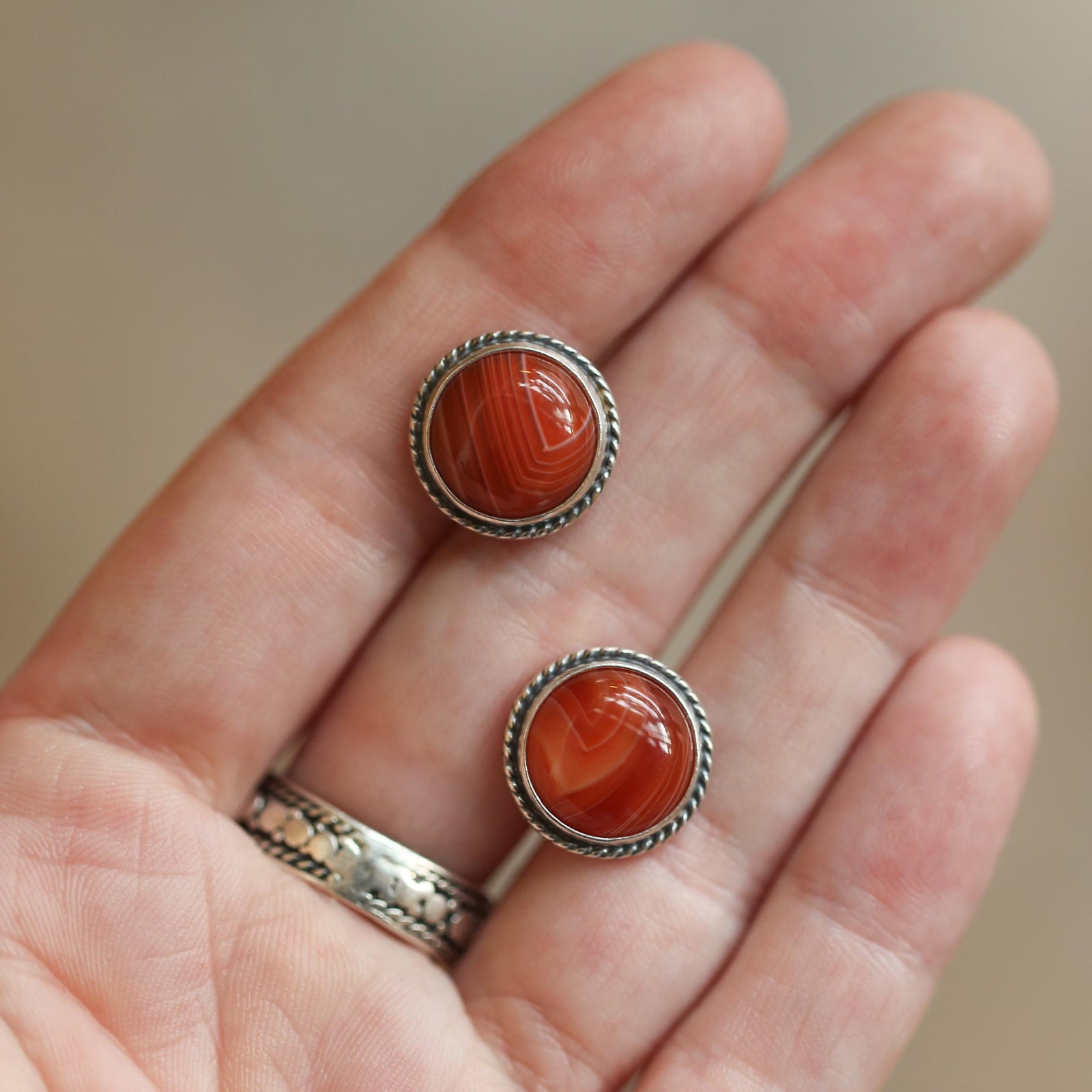 May include: A pair of round stud earrings featuring polished, reddish-brown agate stones with a striped pattern. The stones are set in a silver-colored metal with a twisted rope design around the edge. The earrings are displayed on a hand.