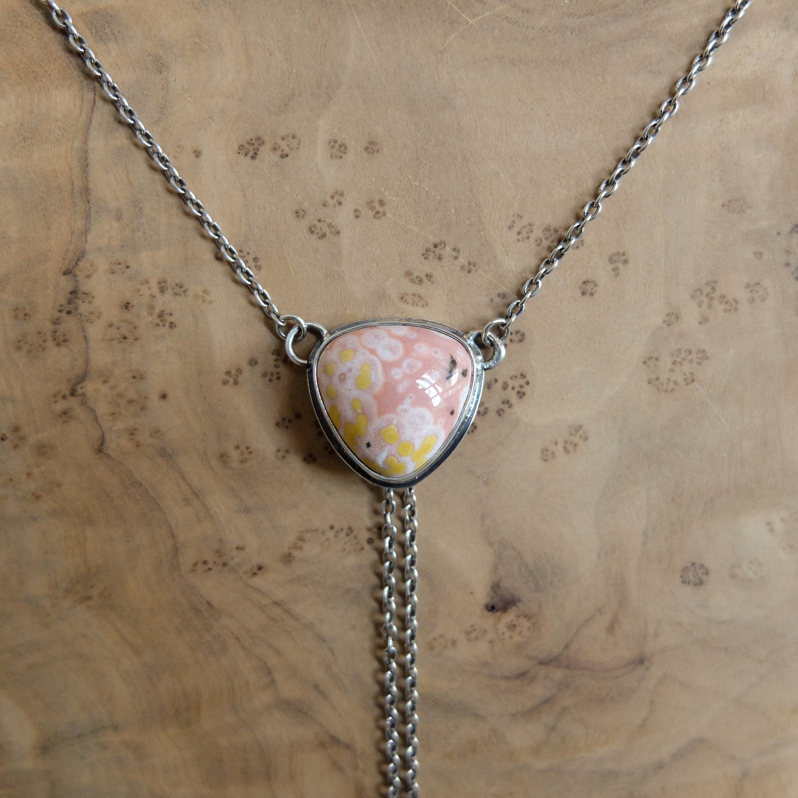 May include: A silver necklace featuring a triangular pendant with a pink and yellow stone. The stone has a unique pattern and is set in a silver bezel. The necklace chain is delicate, and a silver chain dangles from the bottom of the pendant.