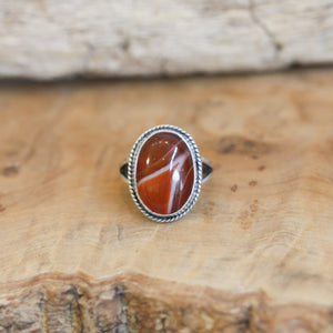 May include: A silver ring with an oval shaped red and white agate stone set in the center. The ring has a rope-like design around the edge of the setting.
