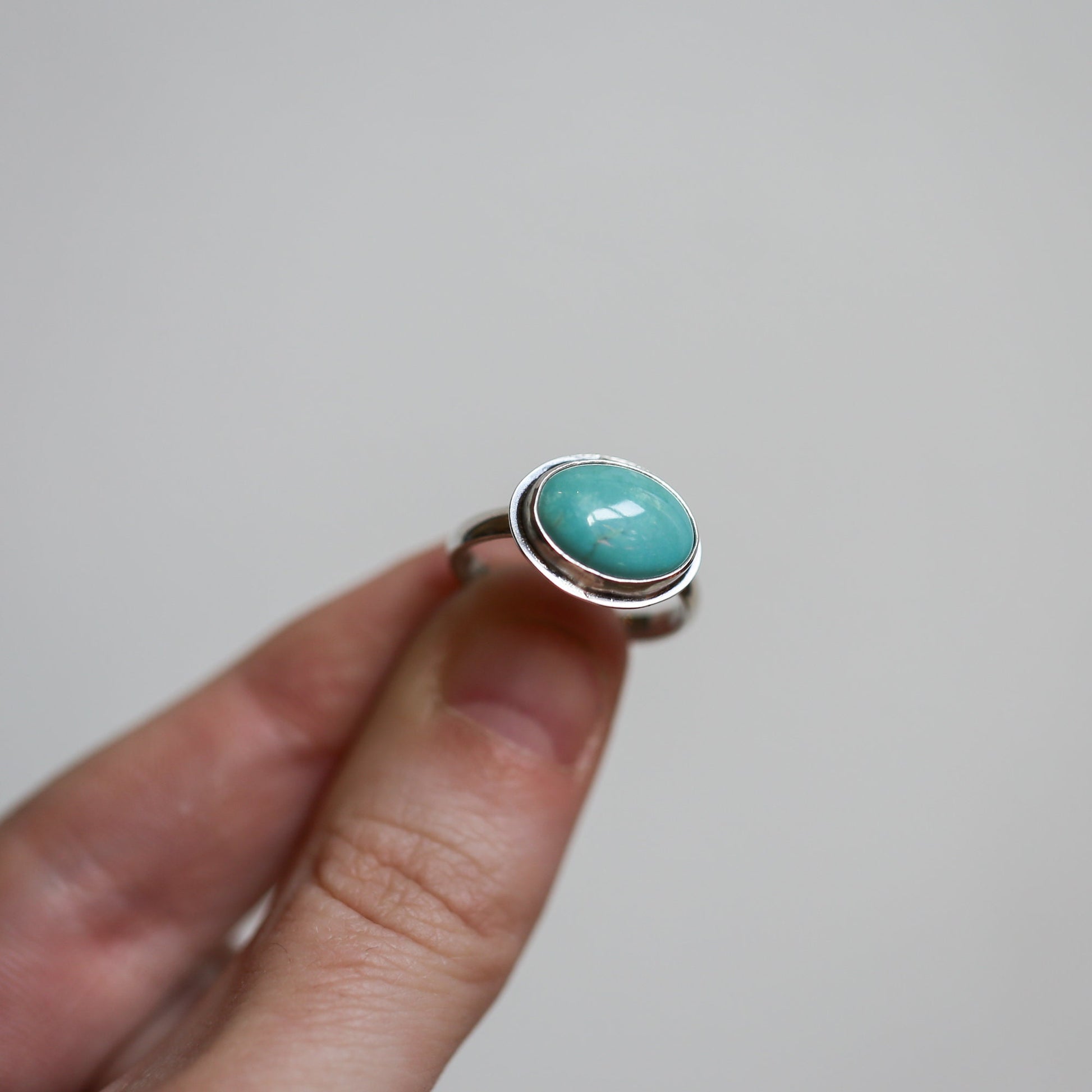 May include: A silver ring with a turquoise gemstone set in a bezel setting. The ring is on a finger.