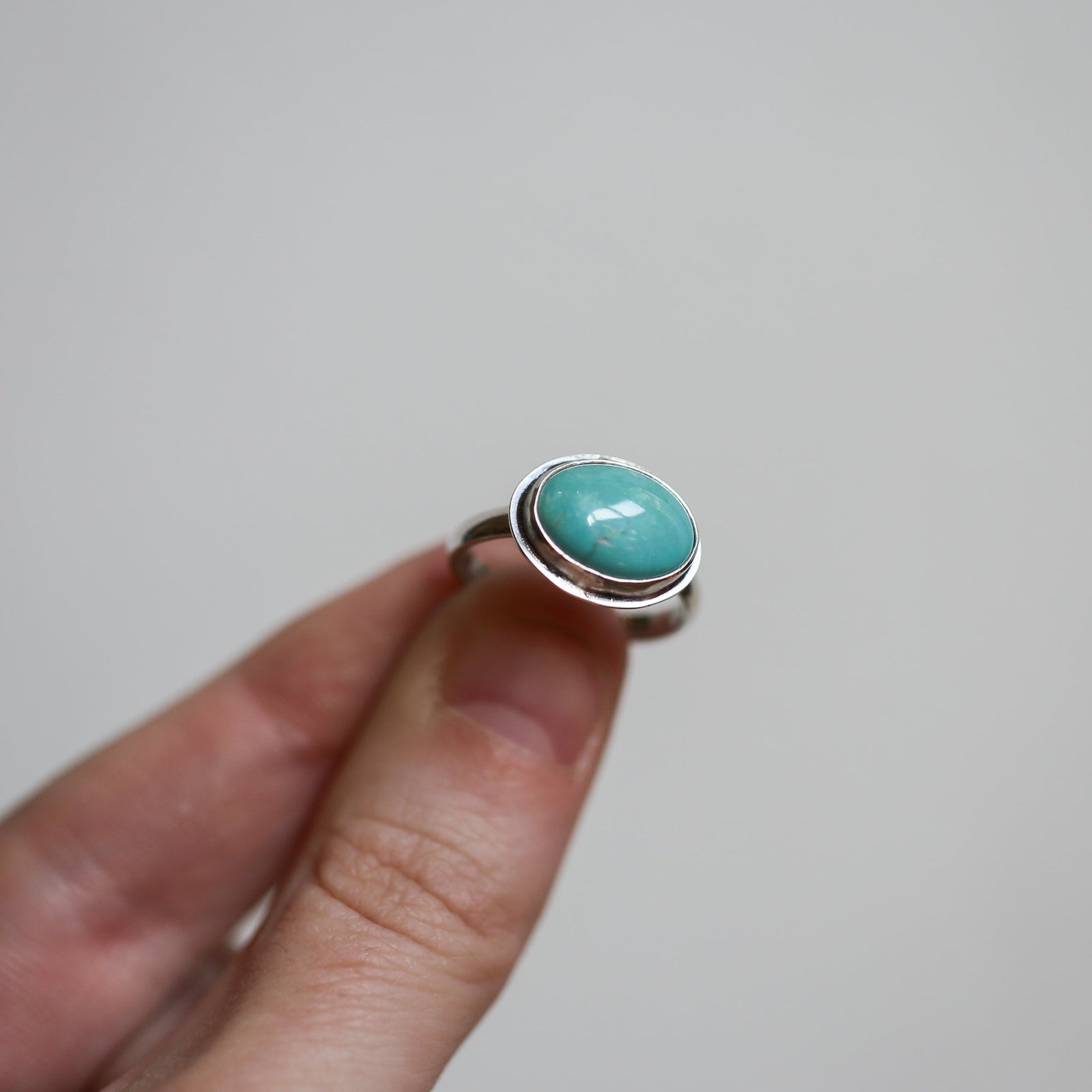 May include: A silver ring with a turquoise gemstone set in a bezel setting. The ring is on a finger.