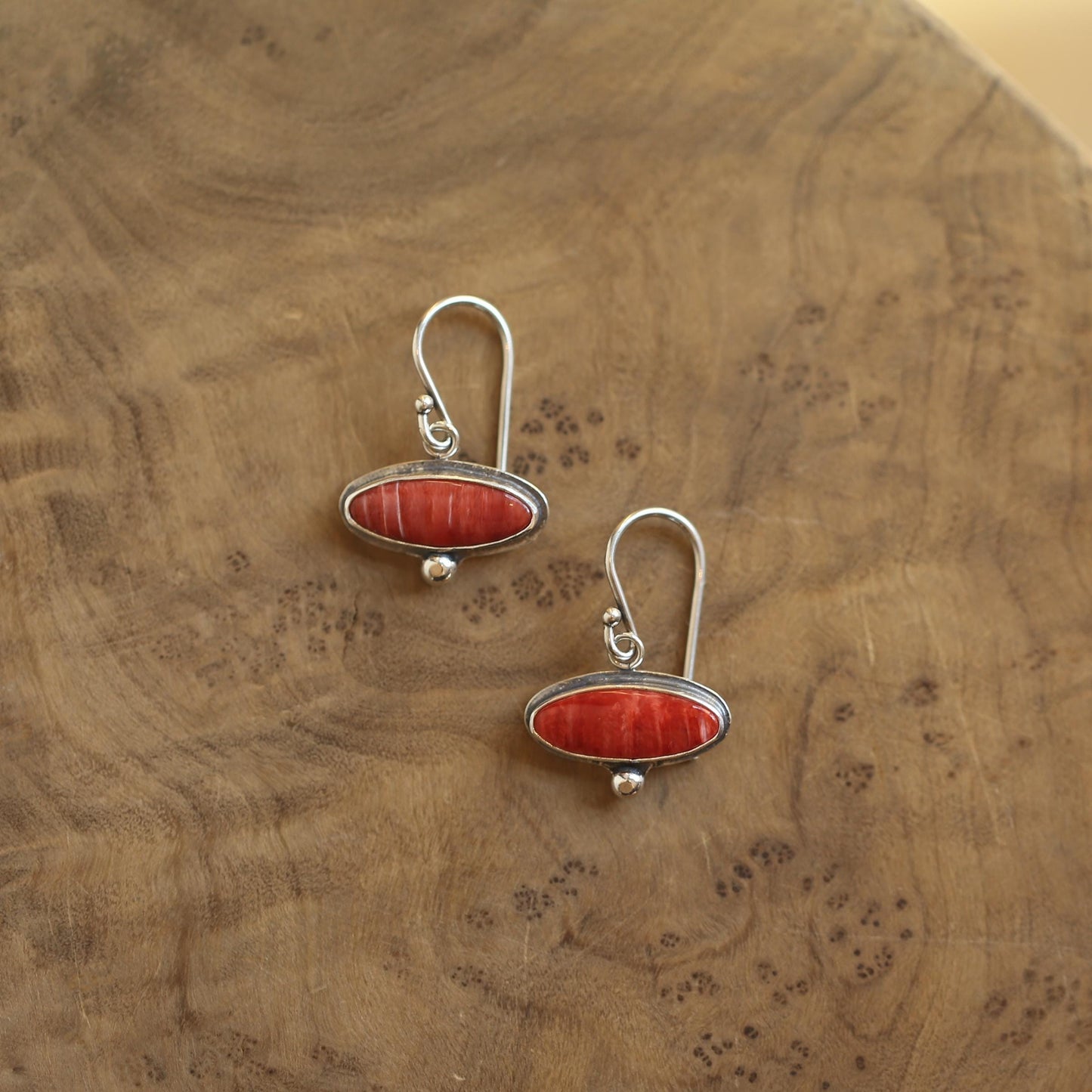 May include: A pair of silver earrings with a red oval-shaped stone. The earrings have a hook closure and a small silver ball at the bottom of the oval. The earrings are displayed on a wooden surface.