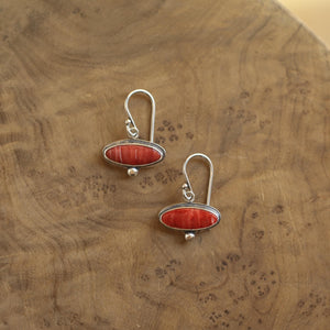 May include: A pair of silver earrings with a red oval-shaped stone. The earrings have a hook closure and a small silver ball at the bottom of the oval. The earrings are displayed on a wooden surface.