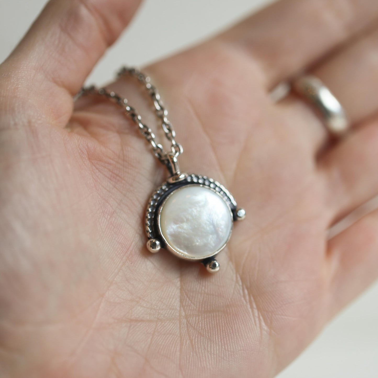 May include: A silver necklace with a round white mother-of-pearl pendant. The pendant is surrounded by a silver frame with a beaded edge.
