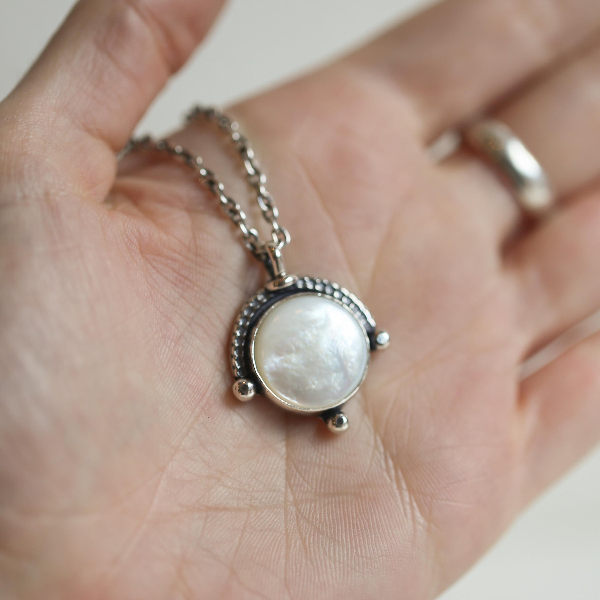 May include: A silver necklace with a round white mother-of-pearl pendant. The pendant is surrounded by a silver frame with a beaded edge.