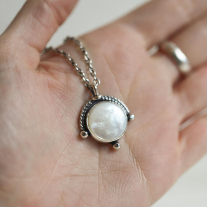 May include: A silver necklace with a round white mother-of-pearl pendant. The pendant is surrounded by a silver frame with a beaded edge.