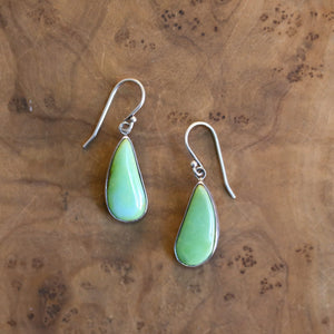 May include: A pair of teardrop-shaped earrings with a light green stone set in a silver metal frame. The earrings have a simple hook design. The background is a warm, brown wooden surface.