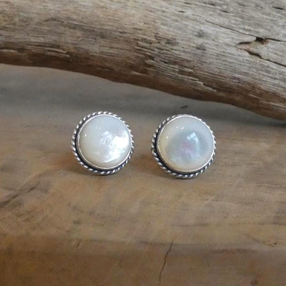 May include: A pair of round stud earrings with a mother-of-pearl center and a silver-colored, twisted rope border. The earrings are displayed on a wooden surface, with a piece of driftwood in the background.